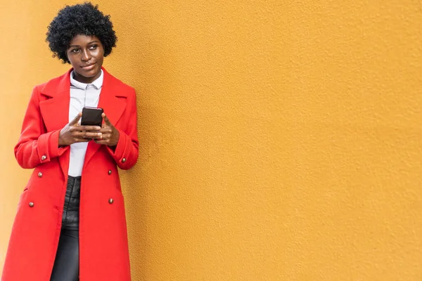 African woman holding a mobile phone while leaning on a yellow wall outdoors on the street. — Stock Photo, Image