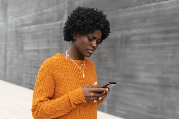Serious black woman looking confused while using a mobile phone outdoors on the street.