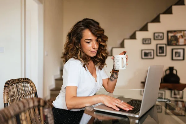 Mujer bebiendo una taza de café mientras trabaja en una computadora portátil en casa. —  Fotos de Stock