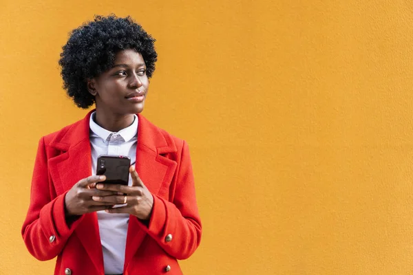 Mulher afro-americana olhando para longe enquanto segurava um telefone celular em um fundo amarelo. — Fotografia de Stock