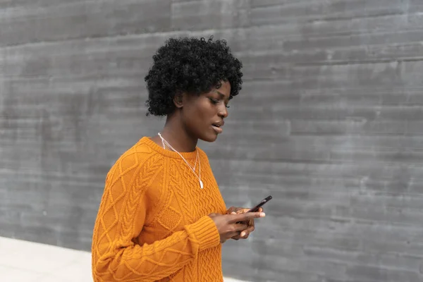 Νεαρή Αφρο γυναίκα δακτυλογραφεί στο κινητό της ενώ περπατάει στο δρόμο. — Φωτογραφία Αρχείου