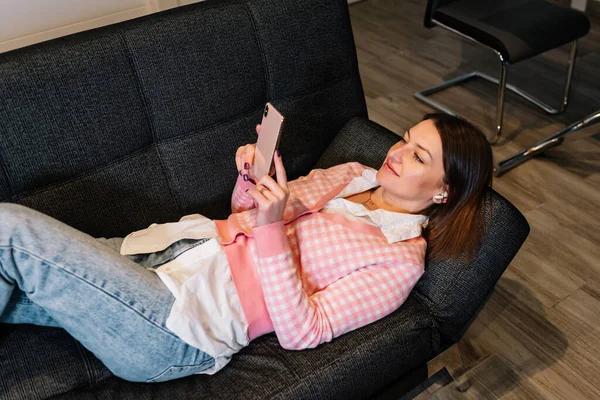 Mujer relajada usando su teléfono móvil mientras está acostada en un cómodo sofá en casa. —  Fotos de Stock