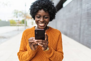 Sokakta cep telefonundan bir şey izlerken gülümseyen genç afro bir kadın..