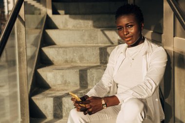 Afrika kökenli Amerikalı kameraya bakıyor ve merdivenlerde otururken cep telefonu tutuyor..