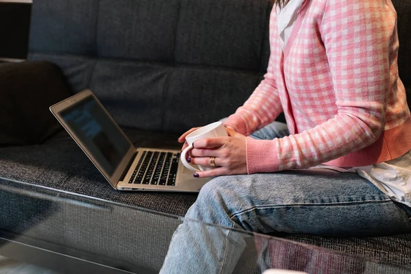 Mujer sosteniendo una taza de café mientras trabaja con un ordenador portátil sentado en el sofá en casa. —  Fotos de Stock