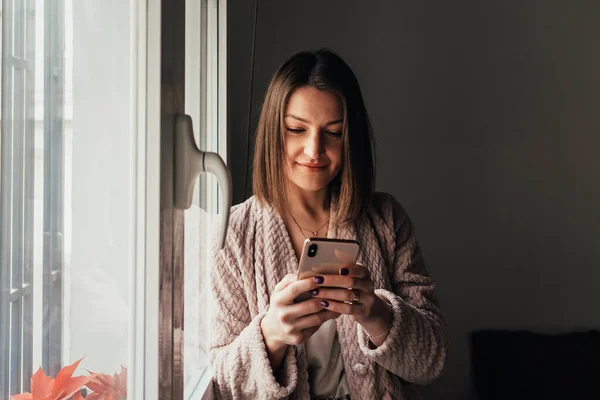 Relaxed woman using her mobile phone while standing next to a window at home. — Stock Photo, Image