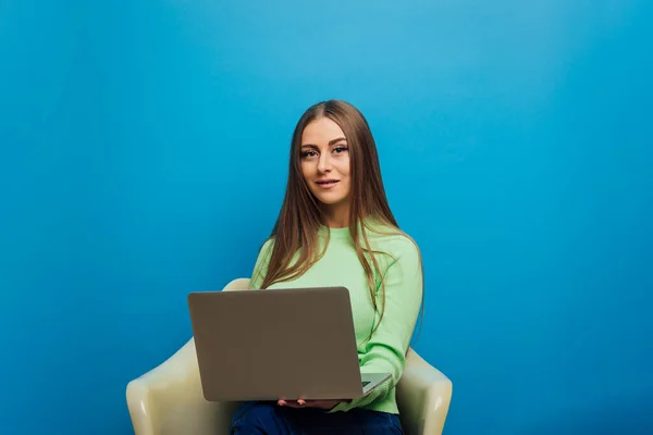 Mujer mirando a la cámara mientras usa una computadora portátil sentada en una silla sobre un fondo aislado. —  Fotos de Stock