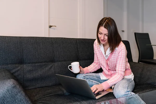 Mujer sosteniendo una taza de café mientras usa un portátil sentado en un sofá en casa. —  Fotos de Stock