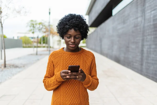 ผู้หญิงสาวชาวแอฟริกันใช้สมาร์ทโฟนของเธอในขณะที่เดินไปตามถนน . — ภาพถ่ายสต็อก