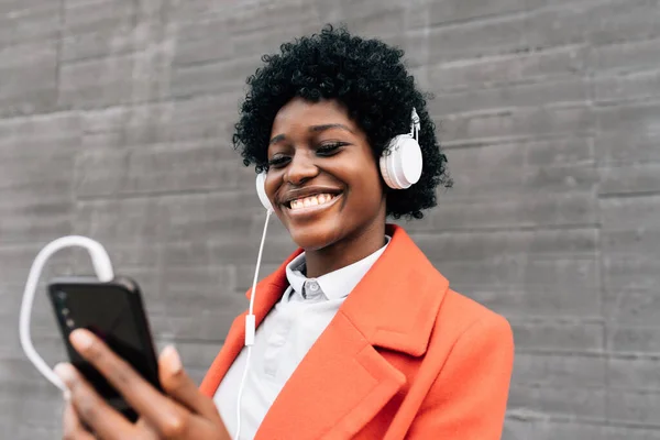 Smiling Afro woman using a mobile phone with headphones while standing outdoors on the street. — Fotografia de Stock