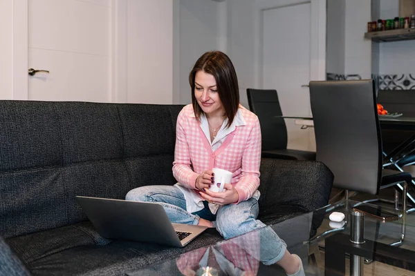 Mujer sosteniendo una taza de café y el uso de una computadora portátil mientras está sentado en un sofá en casa. —  Fotos de Stock