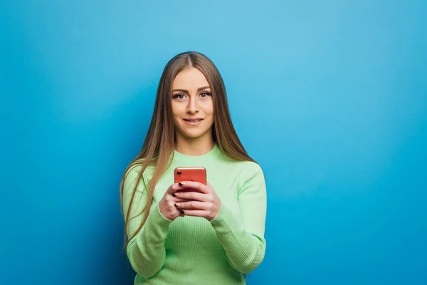 Žena se dívá na kameru, zatímco drží mobilní telefon stojí na izolovaném pozadí. — Stock fotografie