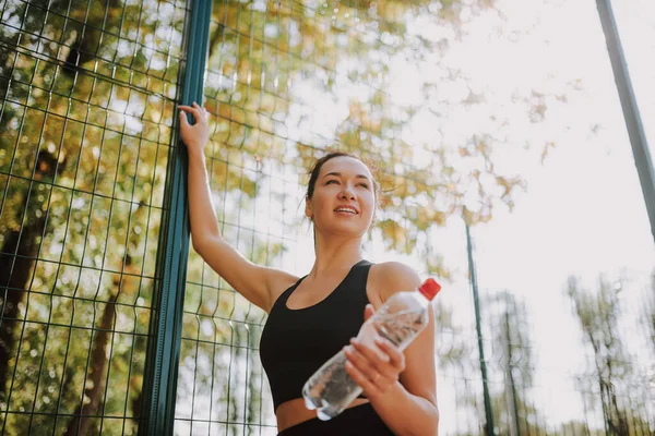 Siyah spor giysili genç bir kadın parkta esniyor ve spor yapıyor. Molada su içen çekici fit bir kız. Sağlıklı yaşam tarzı kavramı.