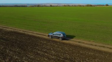 Boş toprak yolda, güneşli bir sabahta tarım arazisi olan yeşil çayırlar boyunca giden arazi aracının hava görüntüsü.
