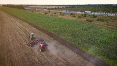 Farmer on tractor with seeding machinery driving over field sows grain of wheat or corn near road with lot of cars at evening