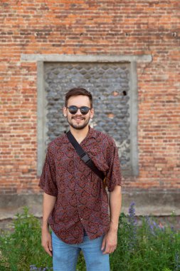 Güneş gözlüklü gülümseyen genç bir hippi dış binanın duvarının yanında duruyor..