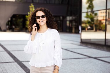 Kıvırcık saçlı, gülümseyen, cep telefonuna bakan genç bir kadın, pencere arkası olan bir binaya bakıyor..