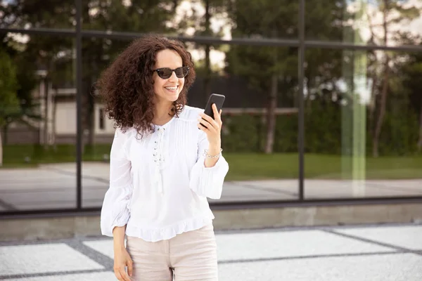 Leende ung kvinna klädd i en vit, tittar på mobilen, mot en byggnad med fönster bakgrund. — Stockfoto