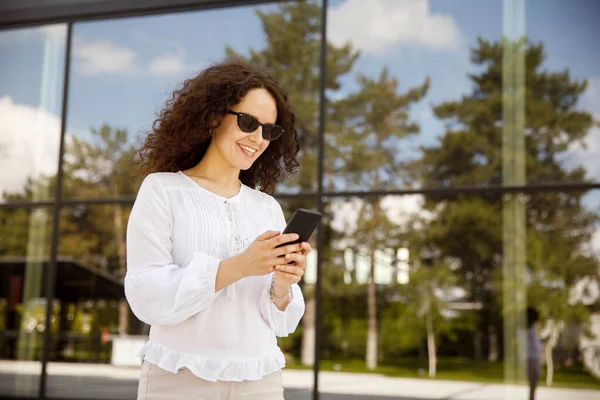 Närbild porträtt av en flicka med lockigt hår, tittar på mobilen, mot en byggnad med fönster bakgrund. — Stockfoto