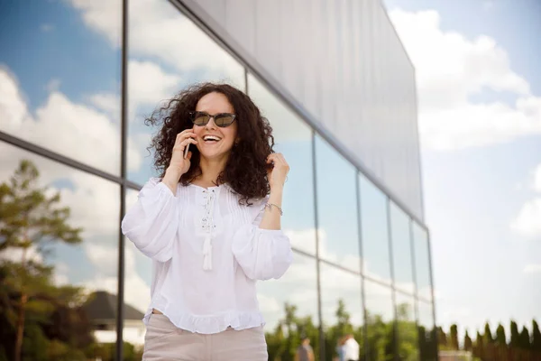 Fröhliches Mädchen mit lockigem Haar, Sonnenbrille, am Handy sprechend, vor einem Gebäude mit Fensterhintergrund. — Stockfoto