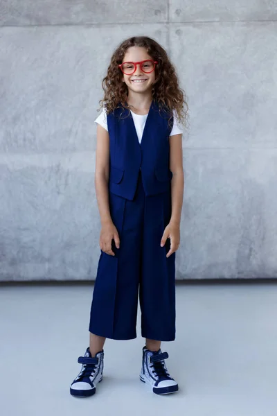 Frontaal portret van een lachend klein meisje in blauw uniform, in rood oogglas, kijkend naar de camera, over een betonnen achtergrond. — Stockfoto