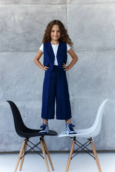 Pretty curly 7s girl sits in feet on a two chairs feeling confident, in a school uniform, over concrete background.