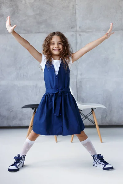 Porträtt av en söt lockig flicka, i klänning poserar i studion med händerna och fötterna i sidled, över konkret bakgrund. — Stockfoto