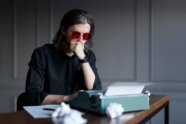 Ung författare i röda glasögon och svart skjorta, tänker på ny bok, arbetar på en skrivmaskin, sitter vid ett bord. — Stockfoto