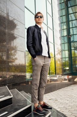 İş merkezinin yanında poz veren, sıradan kıyafetler giyen yakışıklı bir genç resmi..