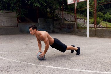 Formda, güçlü, kaslı, üstsüz genç bir adam dışarıda basket topuyla şınav çekiyor. Spor konsepti.