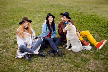 Bir grup genç, gülümseyen insan, köpekleri ile birlikte çimlerin üzerinde oturarak vakit geçiriyorlar..