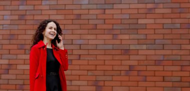 Kırmızılı mutlu kadın, dışarıdaki akıllı telefonda konuşuyor, tuğla duvarın yanında. Metin için boşluk.