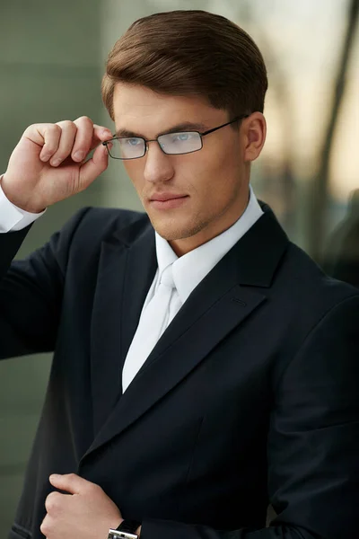 Retrato de un hombre de negocios guapo con gafas, vestido con traje negro y corbata blanca y camisa, sosteniendo sus gafas. — Foto de Stock