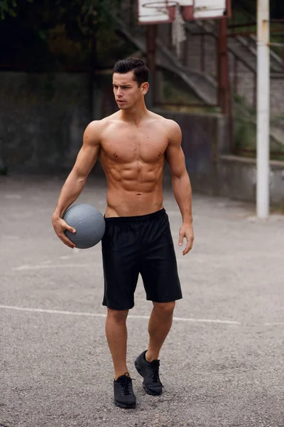 Um jovem forte e musculoso, sem camisa, a segurar uma bola de basquetebol, posa lá fora na quadra urbana. Conceito de desporto. — Fotografia de Stock