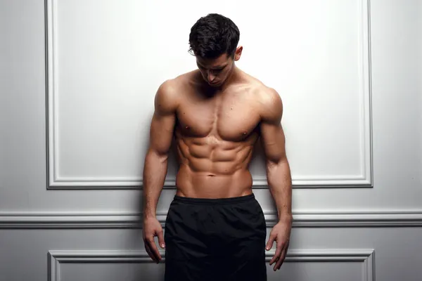 Atleta, hombre musculoso en la pared blanca posa sin camisa, mostrando seis abdominales paquete, fondo blanco. — Foto de Stock