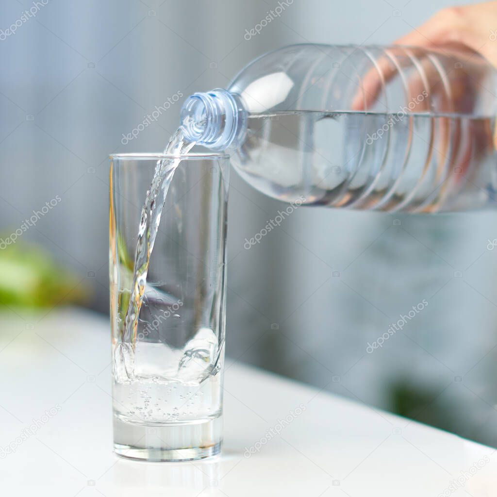 Closeup image of a female hand holding drinking water bottle and pouring water into glass on table on kitchen.