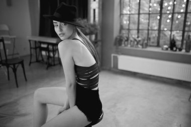 Yatay görüş. Şık giyinmiş, şapkalı, sandalyeye oturmuş kameraya bakan bir kız. Siyah ve beyaz resim.