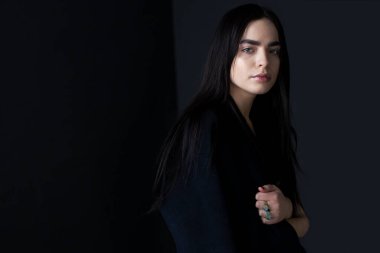 Uzun saçlı, kendine güvenen esmer bir kız, siyah bir ceket giyiyor, stüdyoda poz veriyor, siyah arka planda..