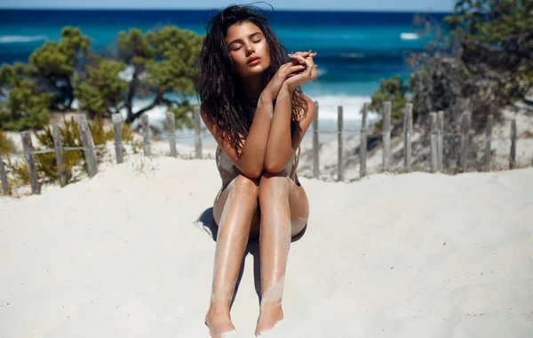 Brunetka seksowna opalona dziewczyna w stroju kąpielowym ciesząca się i siedząca na piasku, z zamkniętymi oczami na plaży, tło krajobrazu morskiego. — Zdjęcie stockowe