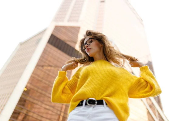 Mulher nova com cabelo ondulado, desgaste em suéter amarelo e jeans brancos sentados atrás do edifício moderno com braço levantado. — Fotografia de Stock