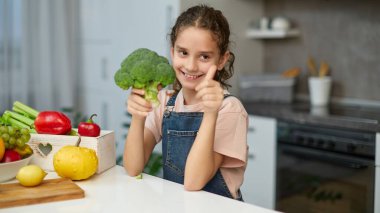 Kıvırcık saçlı küçük bir kız, elinde brokoli, yan tarafa bakıyor, başparmaklarını gösteriyor, mutfaktaki masada oturuyor..