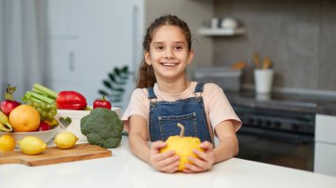 Kıvırcık saçlı, sarı bir balkabağı tutan, mutfaktaki masada oturan tatlı bir kızın ön portresi..
