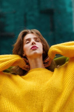 Kahverengi dalgalı saçlı, sarı kazaklı, kendine güvenen genç bir kadın..