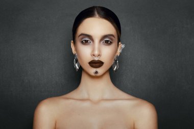 Makyaj mücevherleri olan bir kadın model, kameraya önden bakıyor, stüdyoda poz veriyor, koyu gri arka planda..