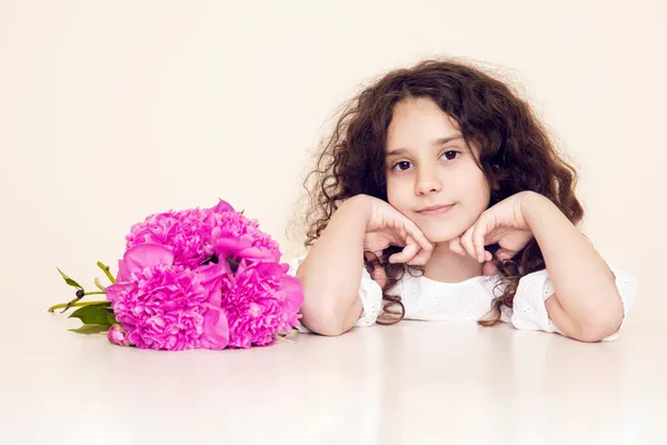 Menina bonita com cabelo encaracolado sentado à mesa, olhando para a câmera, tem um buquê rosa de peônias. — Fotografia de Stock