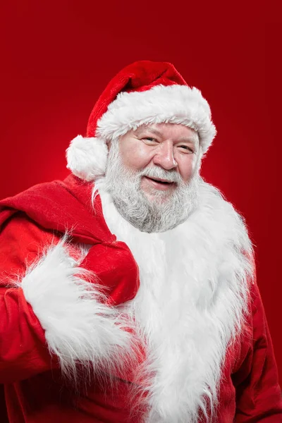 Feliz Santa Claus riendo en un abrigo rojo y un sombrero, mantener una bolsa en la espalda, mirando a la cámara, fondo rojo aislado. —  Fotos de Stock