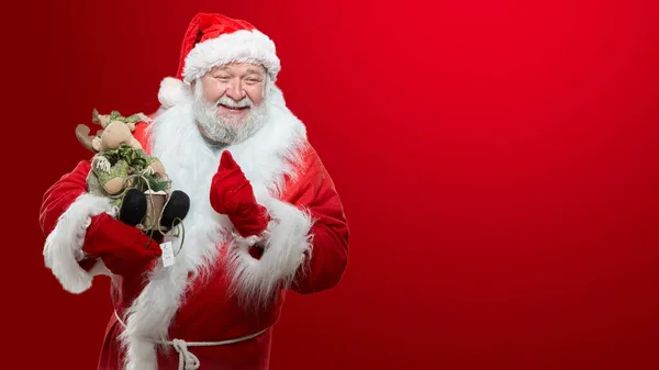 Imagen frontal de un feliz Papá Noel vestido con un abrigo rojo, guantes y un sombrero, sosteniendo un juguete en las manos, señalando su dedo índice, fondo rojo aislado. — Foto de Stock