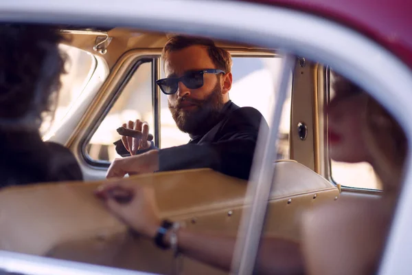 Close up portrait of a stylish bearded man smokes cigar in vintage car. A two men and woman inside of a vintage car.