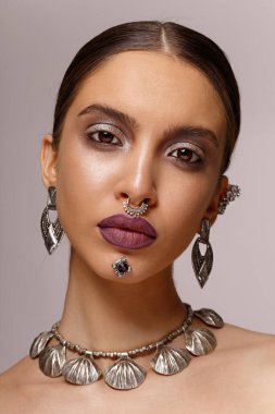 Makyajlı, mücevherli, kulaklı, boyunlu, burunlu, çeneli, gri arka planda kameraya bakan bir kadın modelin yakın plan güzel portresi. Dikey görünüm.