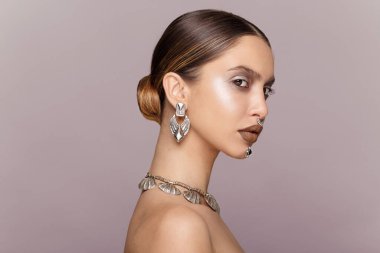 Makyajlı, saç stili, omuzları çıplak, mücevherleri kulakları, boynu, burnu, çenesi gri..